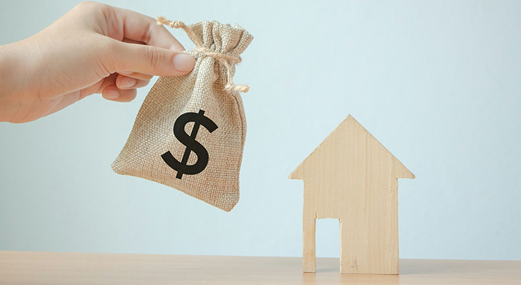 Cropped Hand Of Person Holding Small Sack With Dollar Sign By Model Home On Table